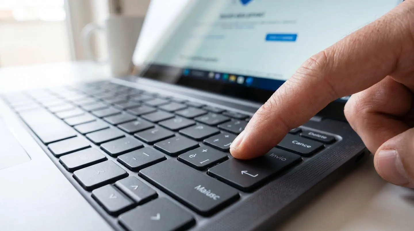 Primo piano estremo di una tastiera di laptop moderna con un dito che preme il tasto Invio, schermo visibile ma completamente sfocato sullo sfondo